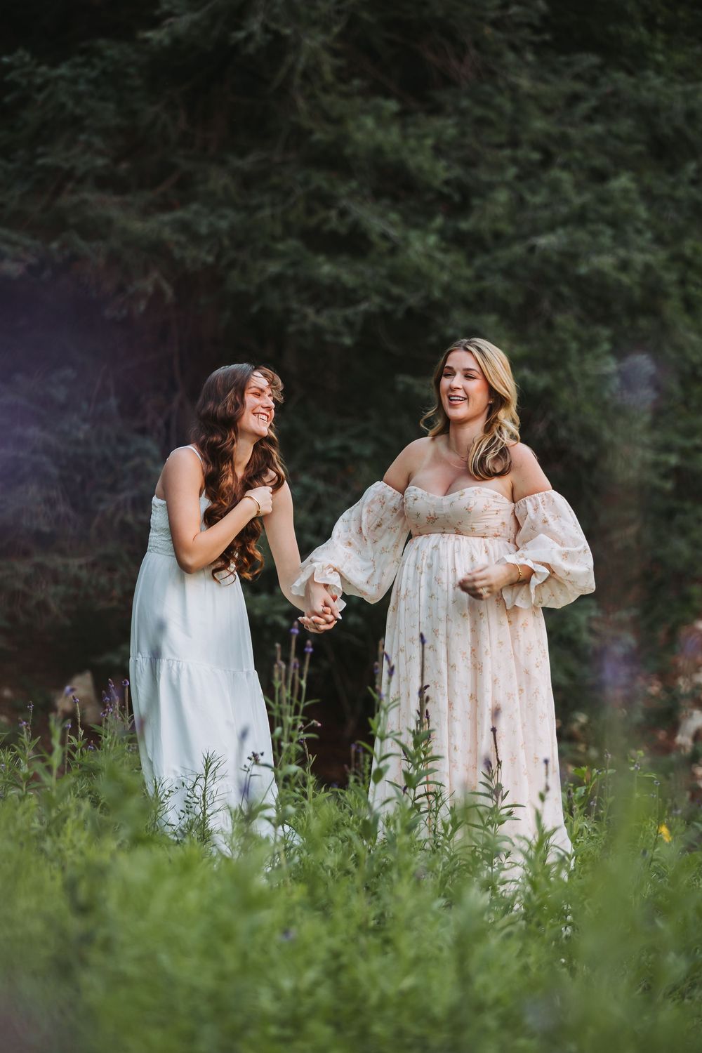 Two friends in white dresses laugh together while standing in a wildflower meadow during a summer photoshoot.