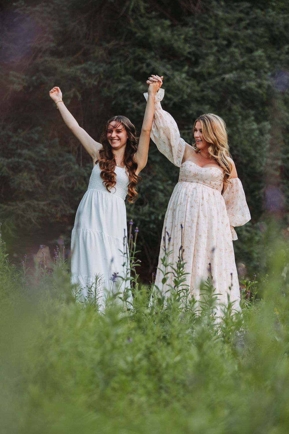 Two women wearing flowy white dresses dance and celebrate together in a lush green meadow on a summer day.