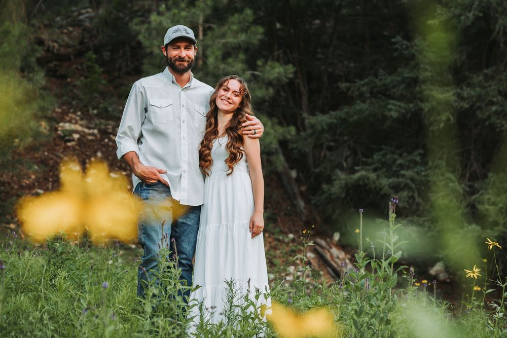 A couple in coordinated white outfits embrace while standing in a lush garden setting during a warm summer day.