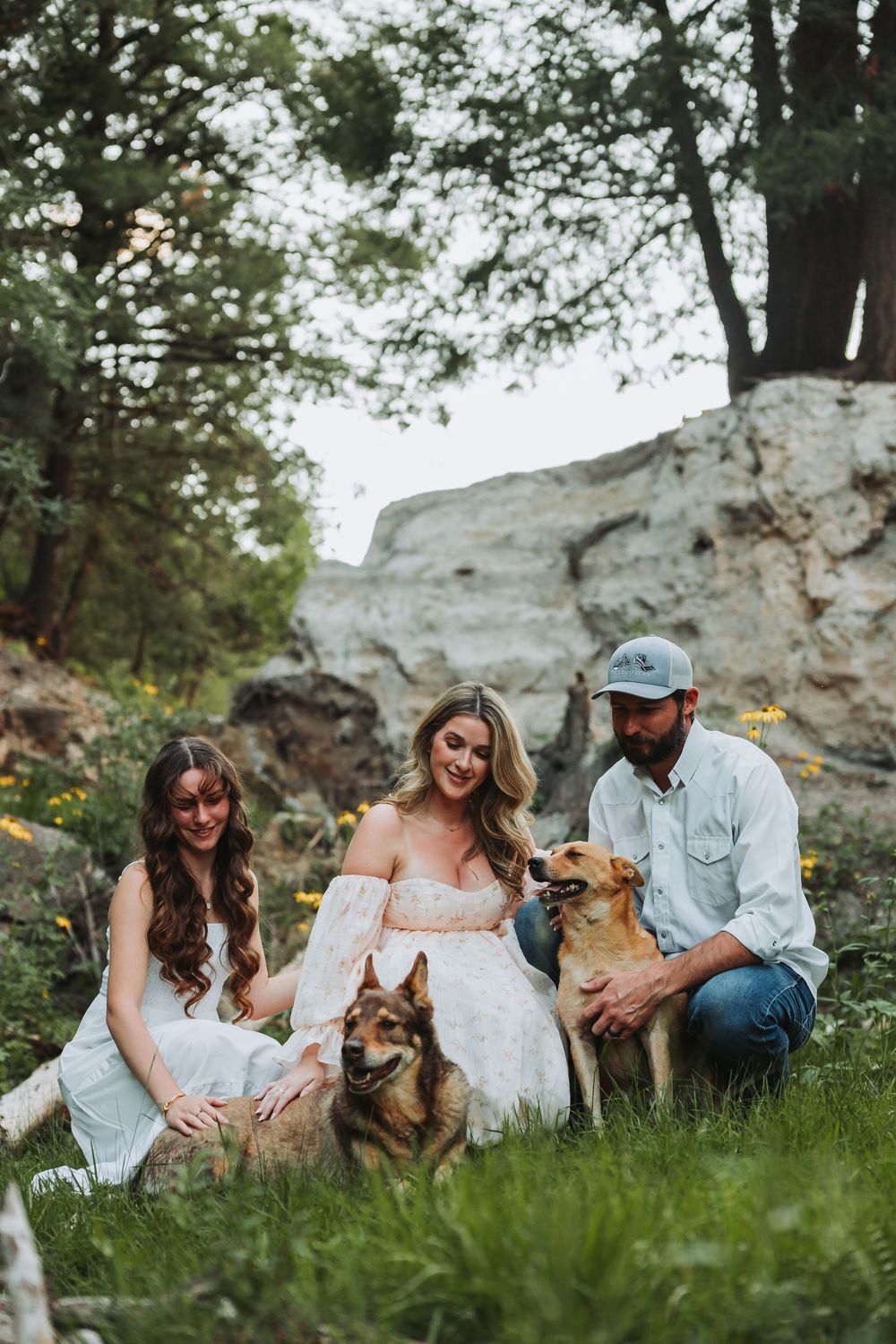 Couple sits in lush grass with two dogs against white rock formation enjoying sunny outdoor photoshoot.