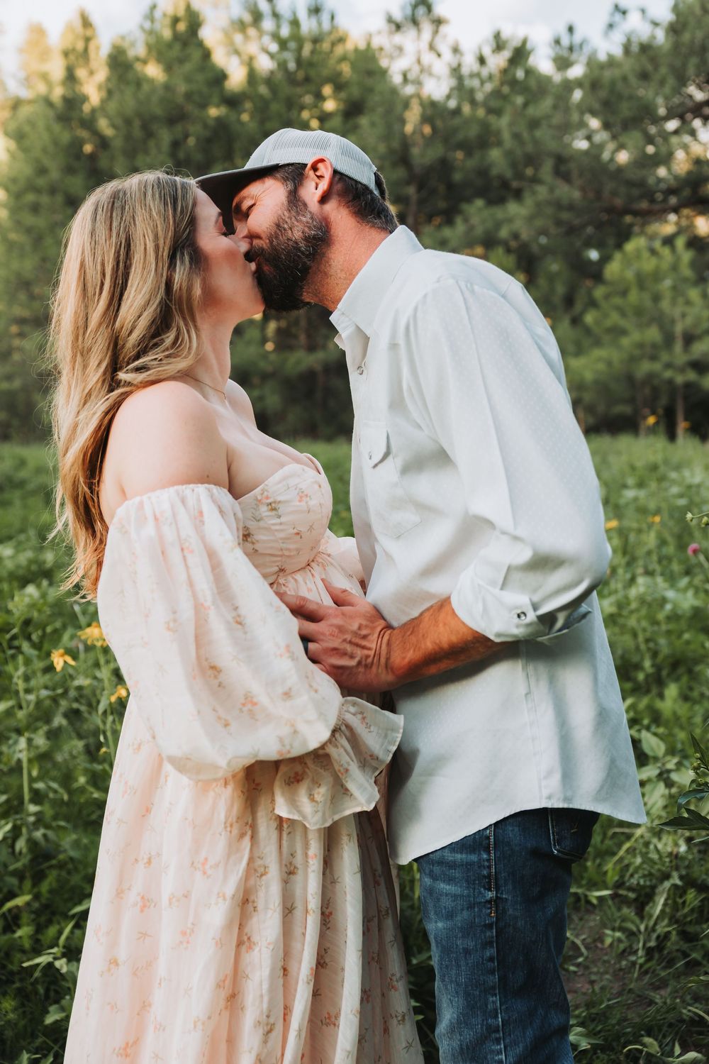 Two people embrace romantically in a sun-drenched field during a photoshoot, surrounded by lush greenery and trees.