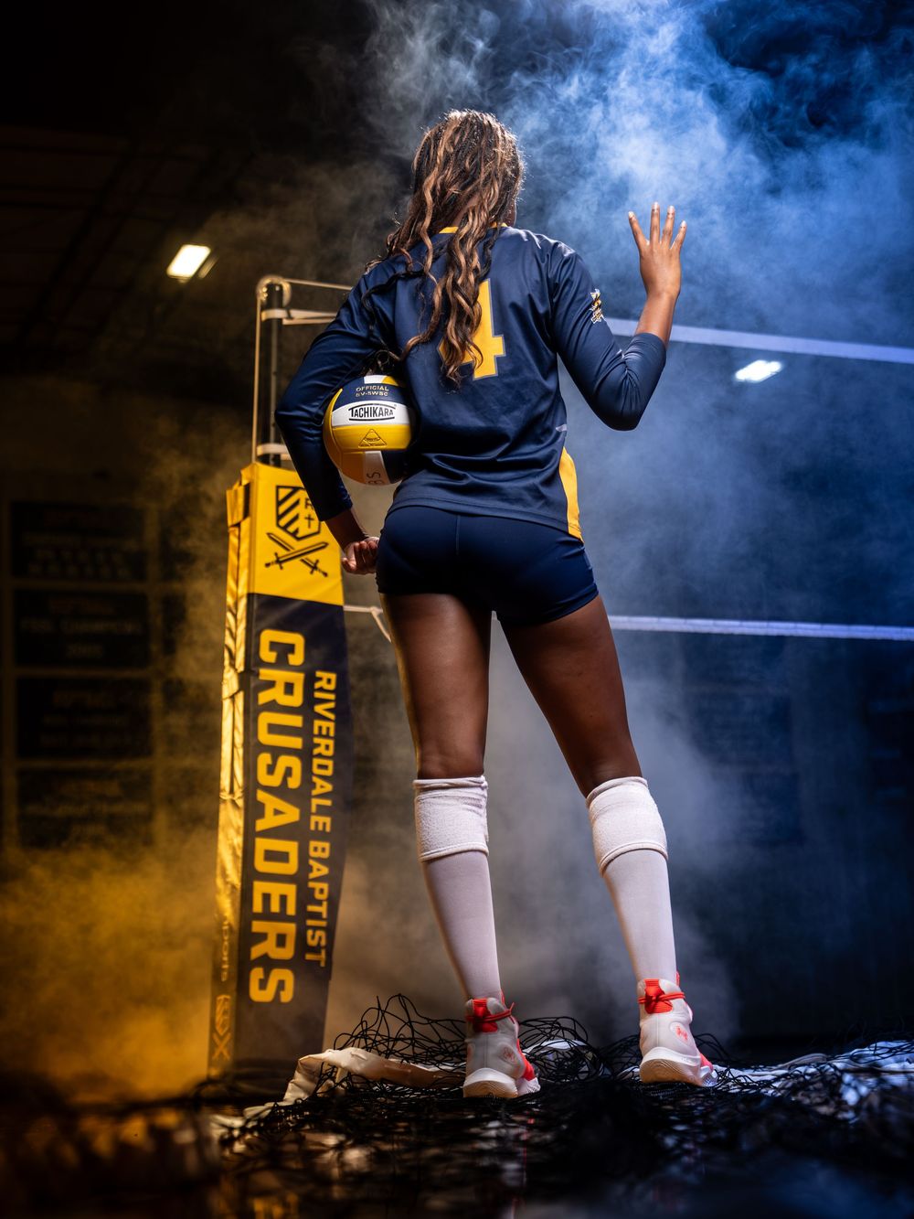 Female Riverdale Baptist Crusaders volleyball player stands at net holding ball in smoky arena.