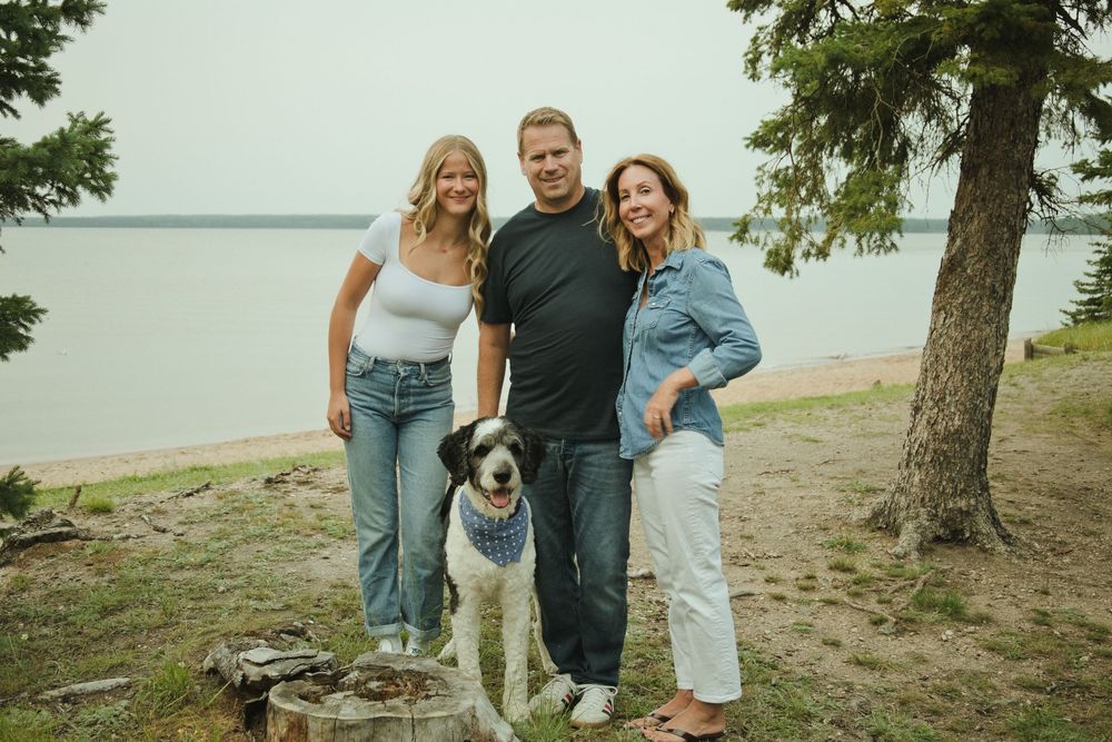 Two women and a man smile with their dog near a lakeside beach surrounded by trees.