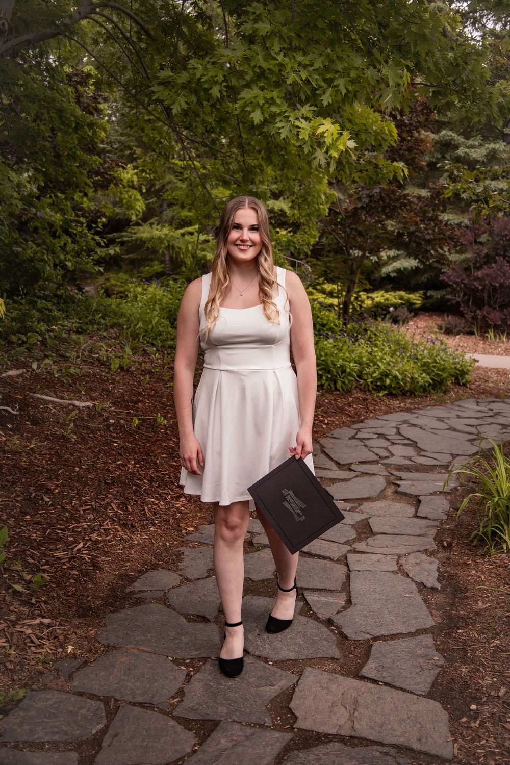 Young woman in white dress holding diploma on stone path in lush garden, celebrating graduation day.