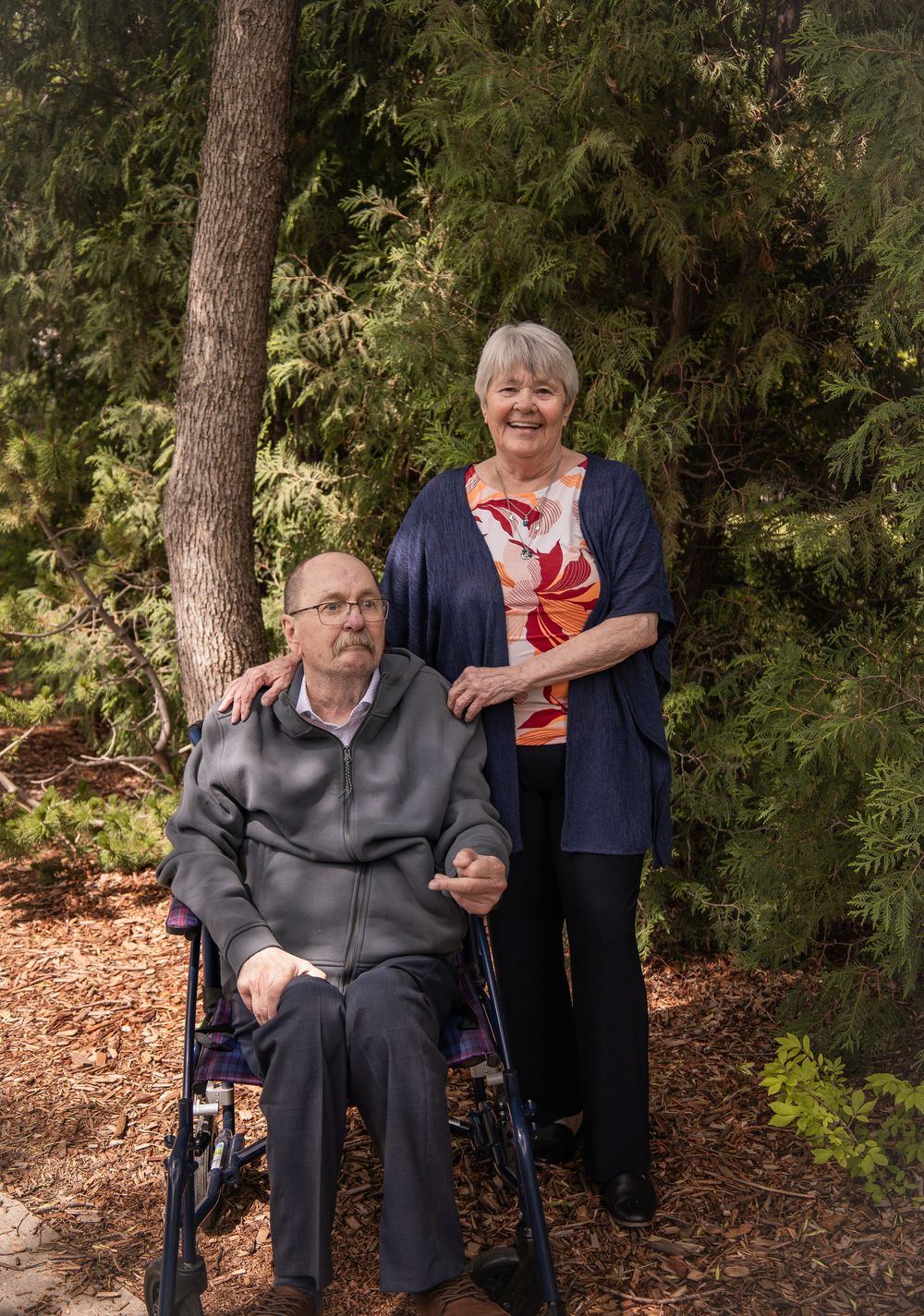 Elderly couple posing outdoors near evergreen trees, man seated in wheelchair, woman standing beside him smiling.