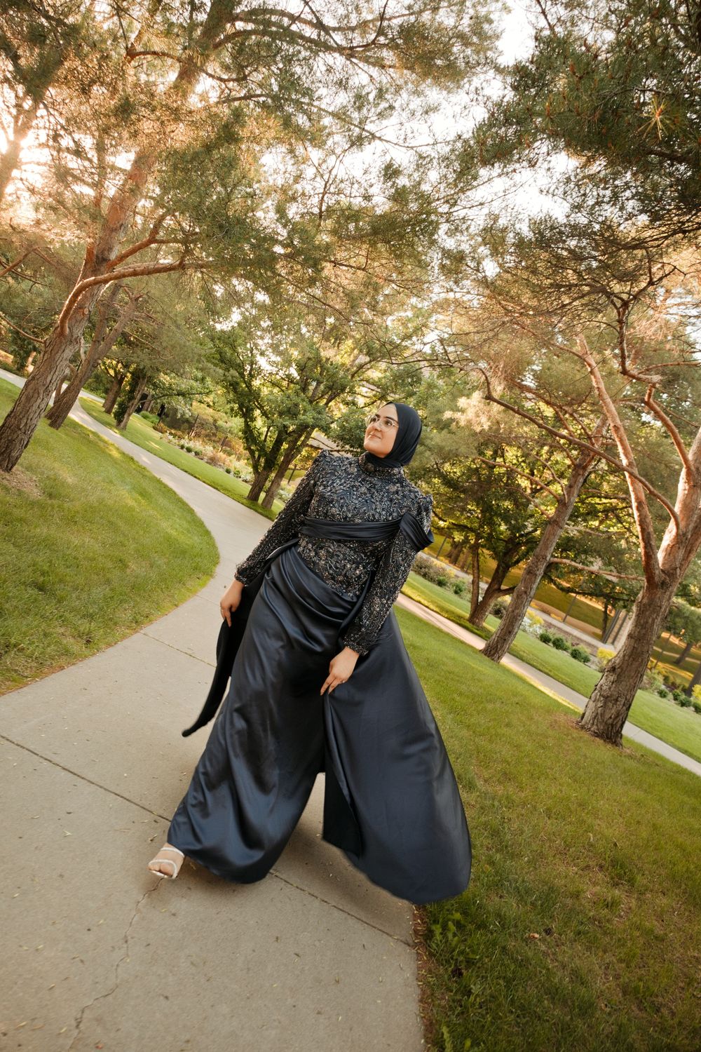 A woman in an elegant black hijab and embellished gown walks gracefully along a park path at sunset.