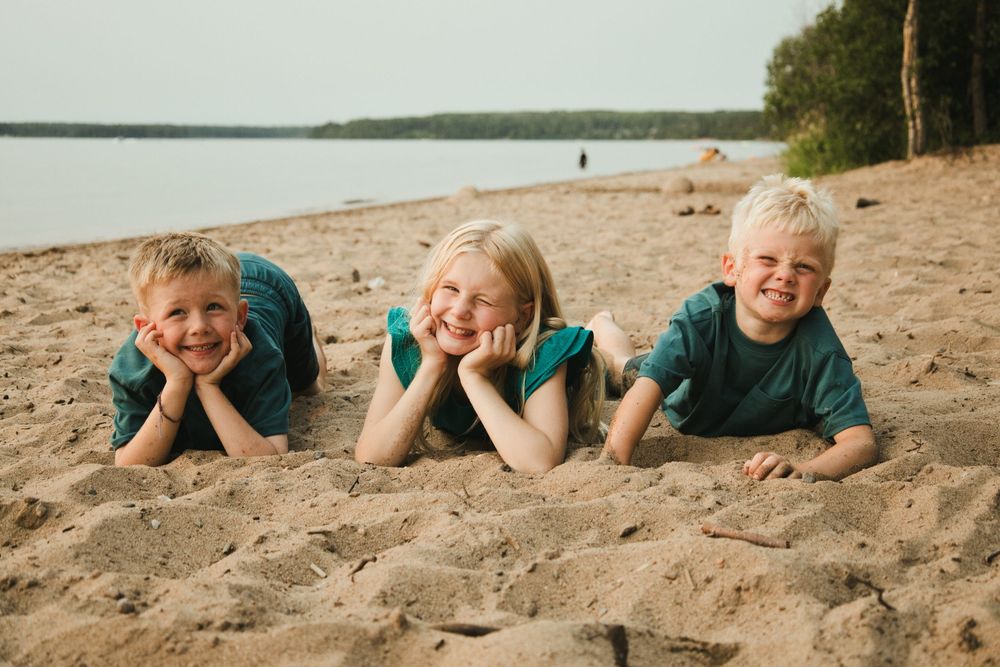 Three smiling children lying on sandy beach near lake, wearing teal shirts, enjoying a sunny summer day outdoors.