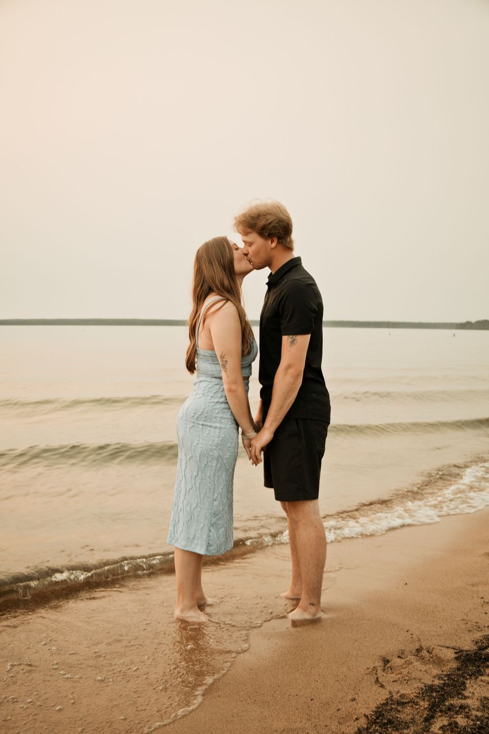 Couple kissing on sandy beach shore, woman in blue dress and man in black outfit, waves gently lapping at their feet.