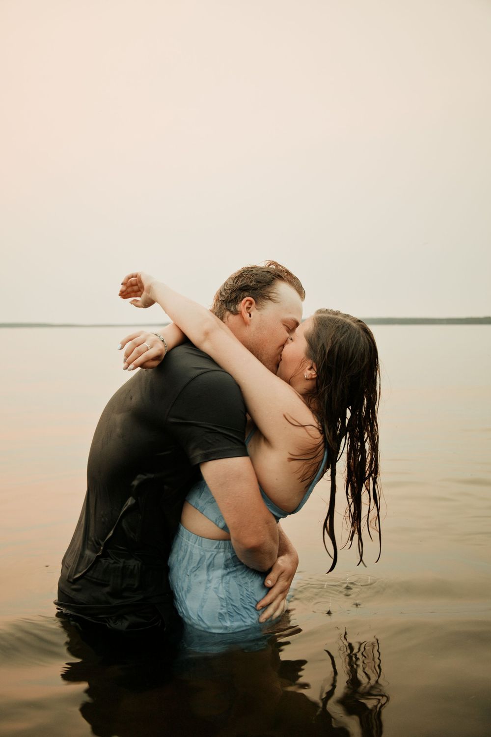 Couple kissing passionately while standing waist-deep in calm lake water during golden hour.