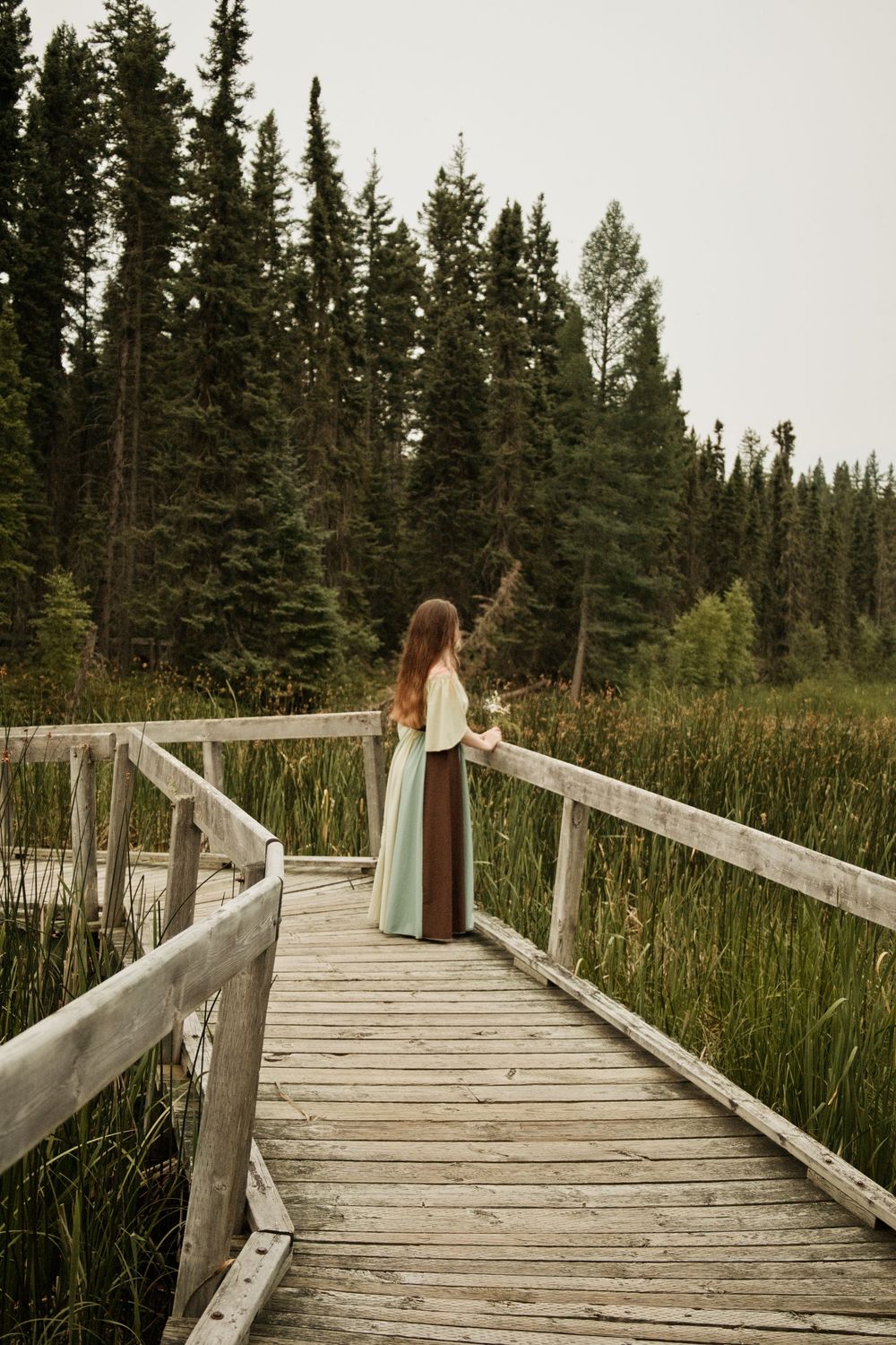 Woman in flowing teal skirt stands on rustic wooden boardwalk surrounded by tall pine trees and marshland.
