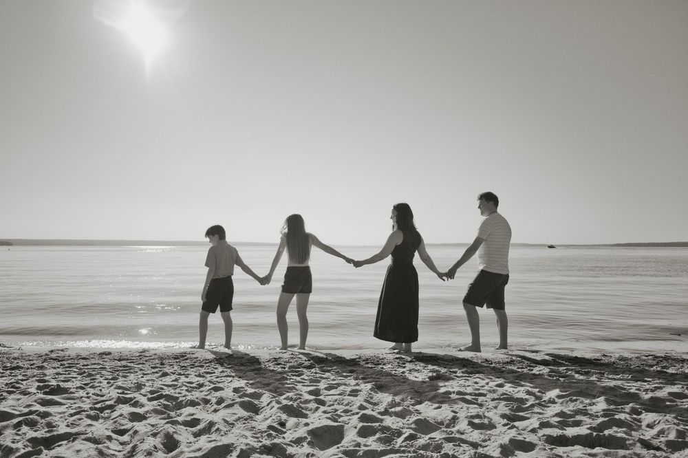 Black and white photo of four people holding hands in a line on a sunny beach near the ocean.