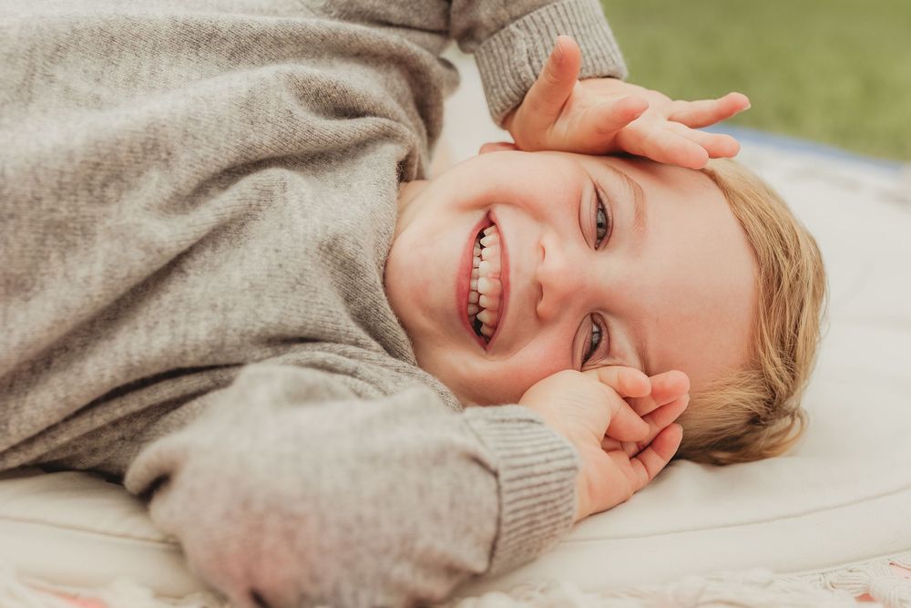 Toddler in gray sweater lying down on white surface with playful expressions.