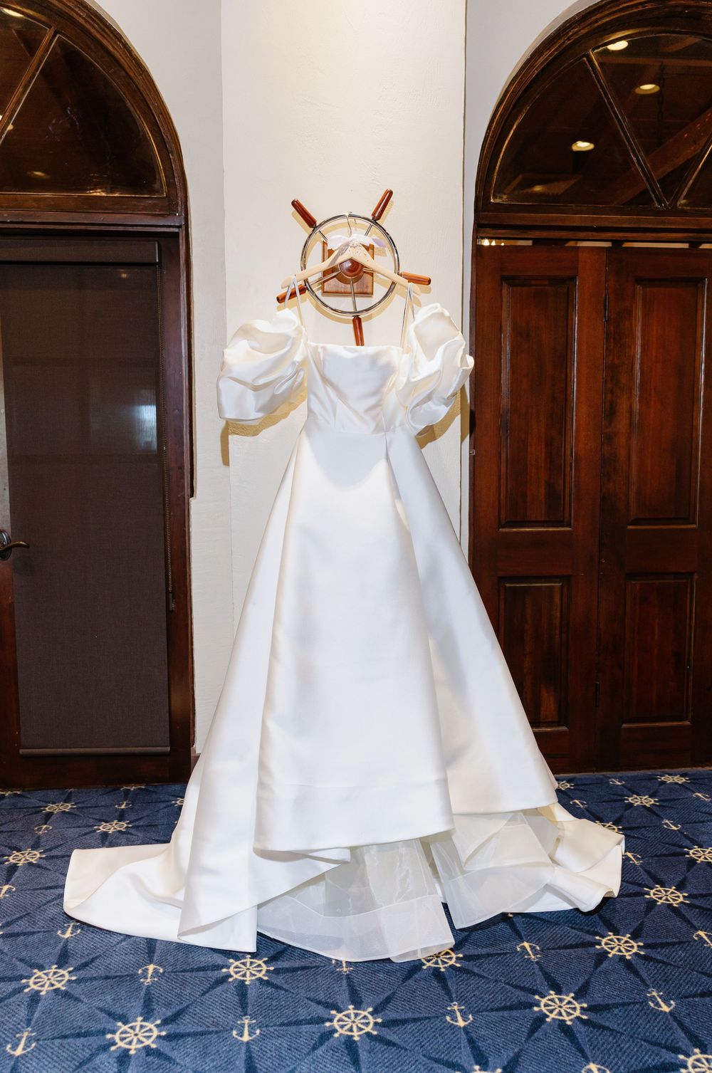 Elegant white wedding dress displayed with nautical ship wheel decor against wooden doors.