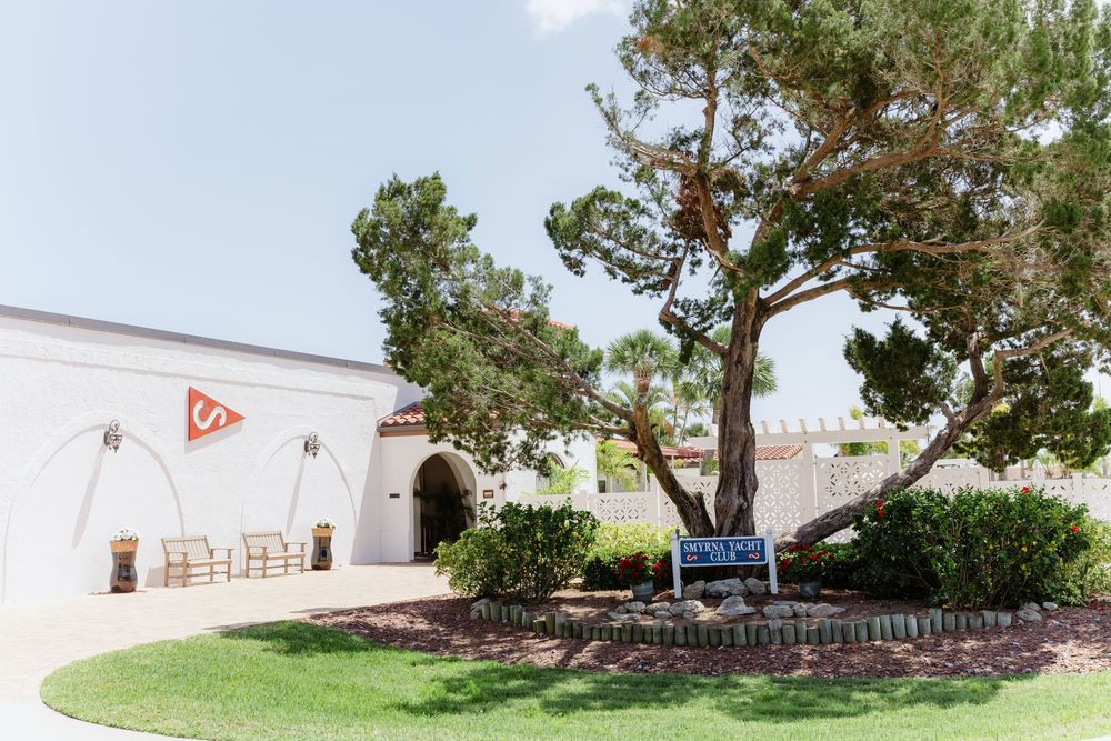 Pine trees line a landscaped area with stone retaining wall and white building in the background.