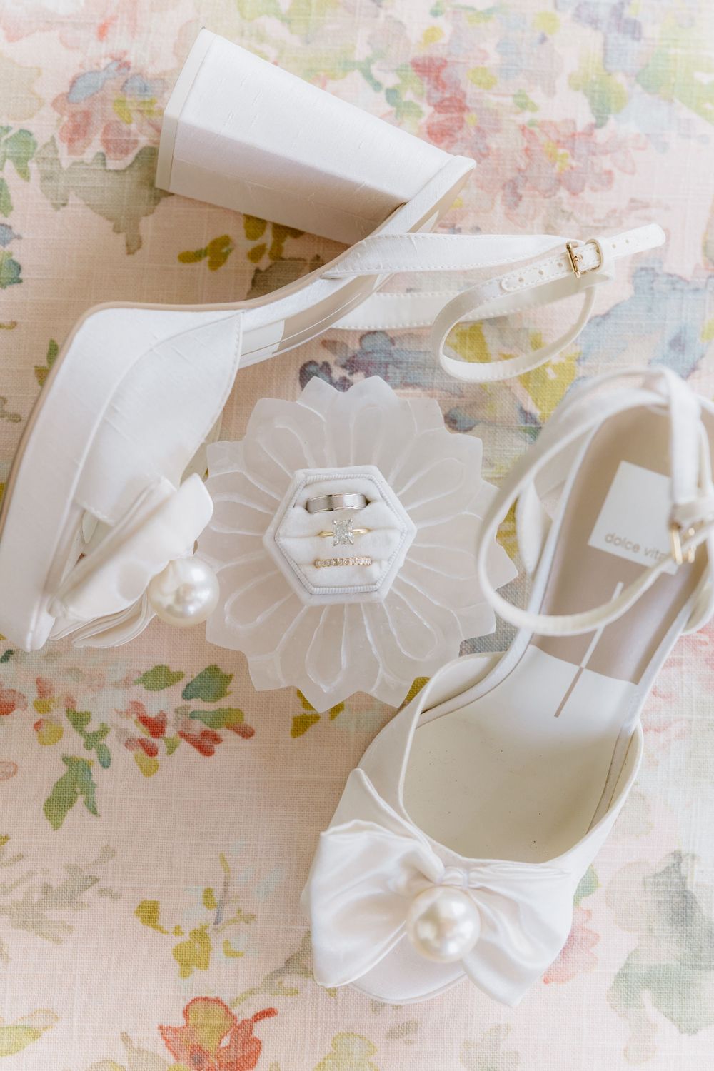 White bridal shoes displayed on delicate floral-patterned wallpaper with feminine design elements.