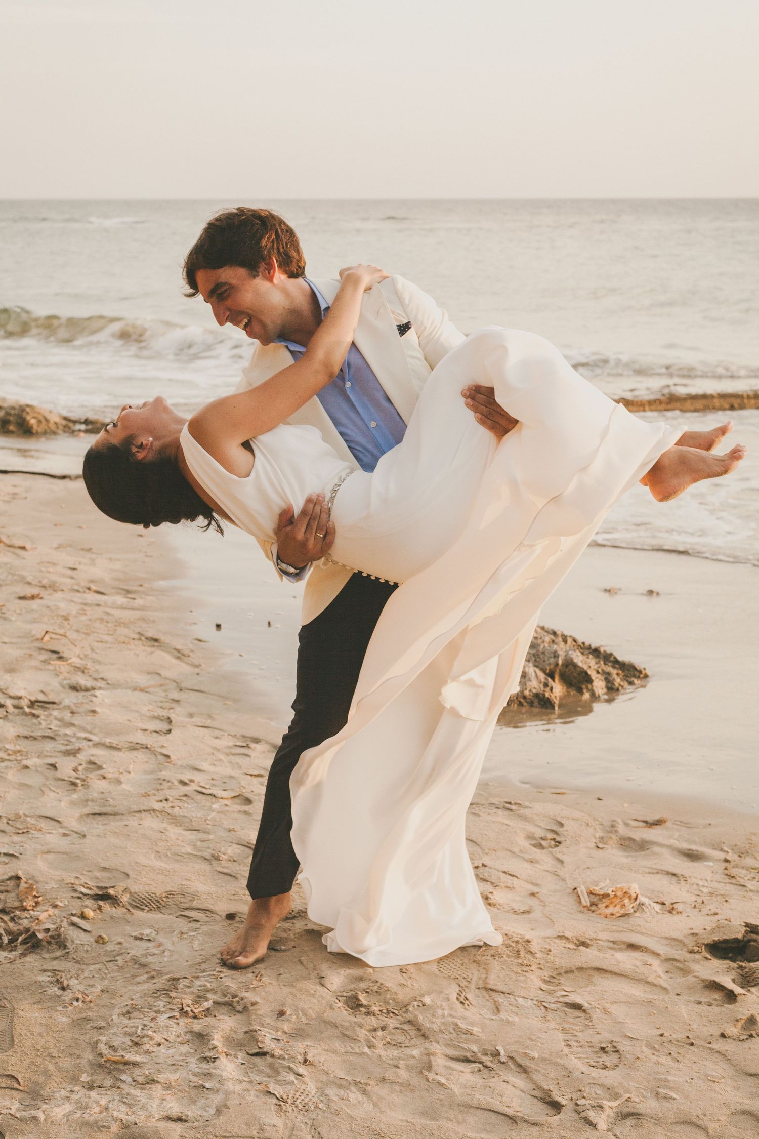 A romantic couple embraces on the beach during a wedding photo session with flowing white dress and dramatic poses.