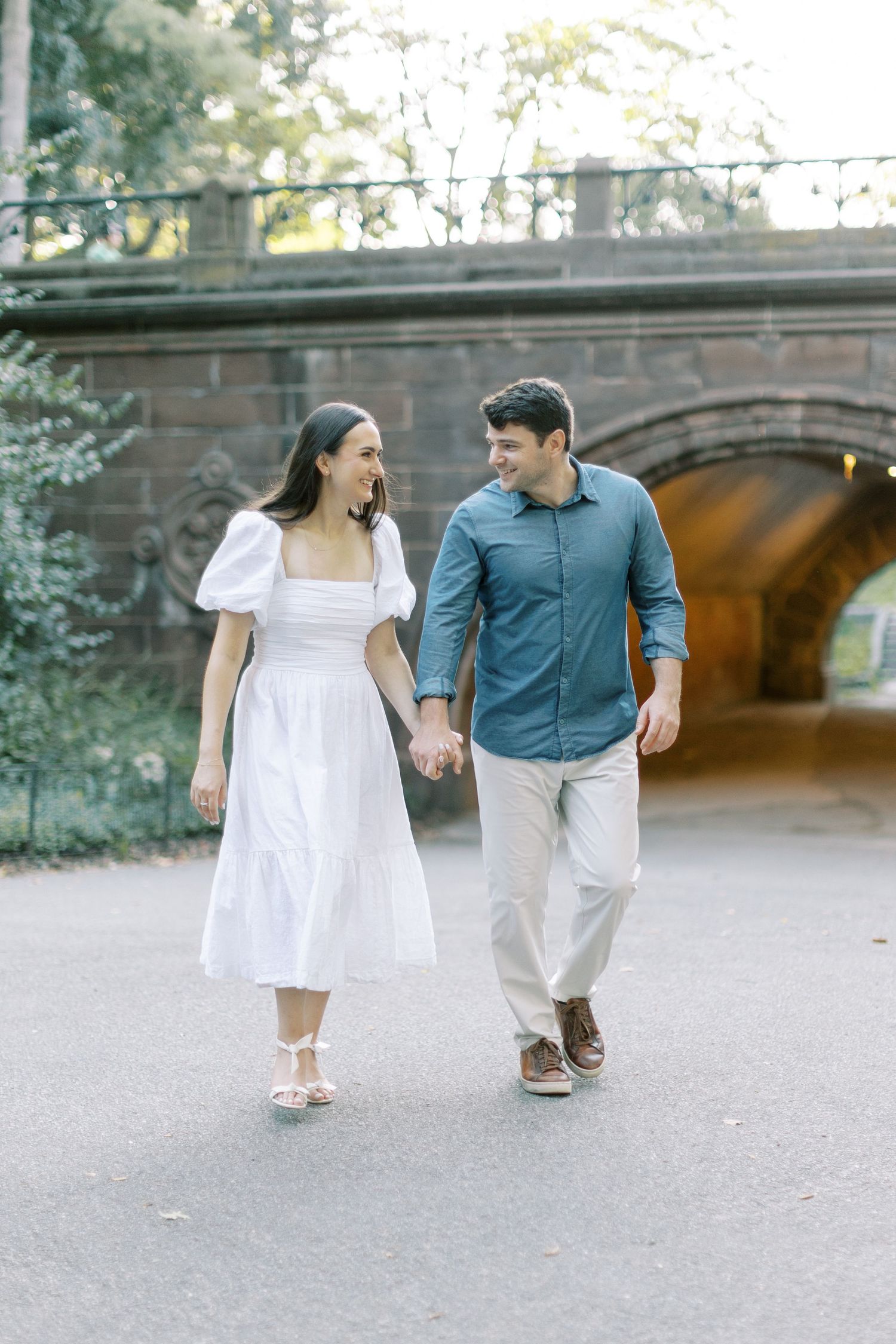 A couple holds hands while walking together near a stone archway tunnel, with the woman in white and man in blue.