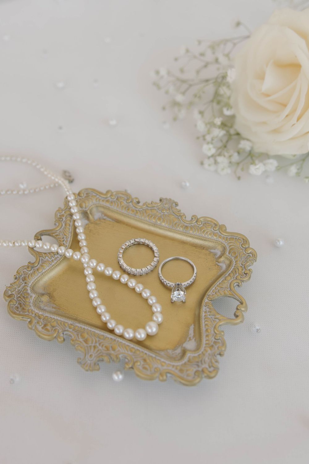 Wedding accessories including pearl necklace and engagement ring displayed on an ornate gold tray with roses and floral decor.