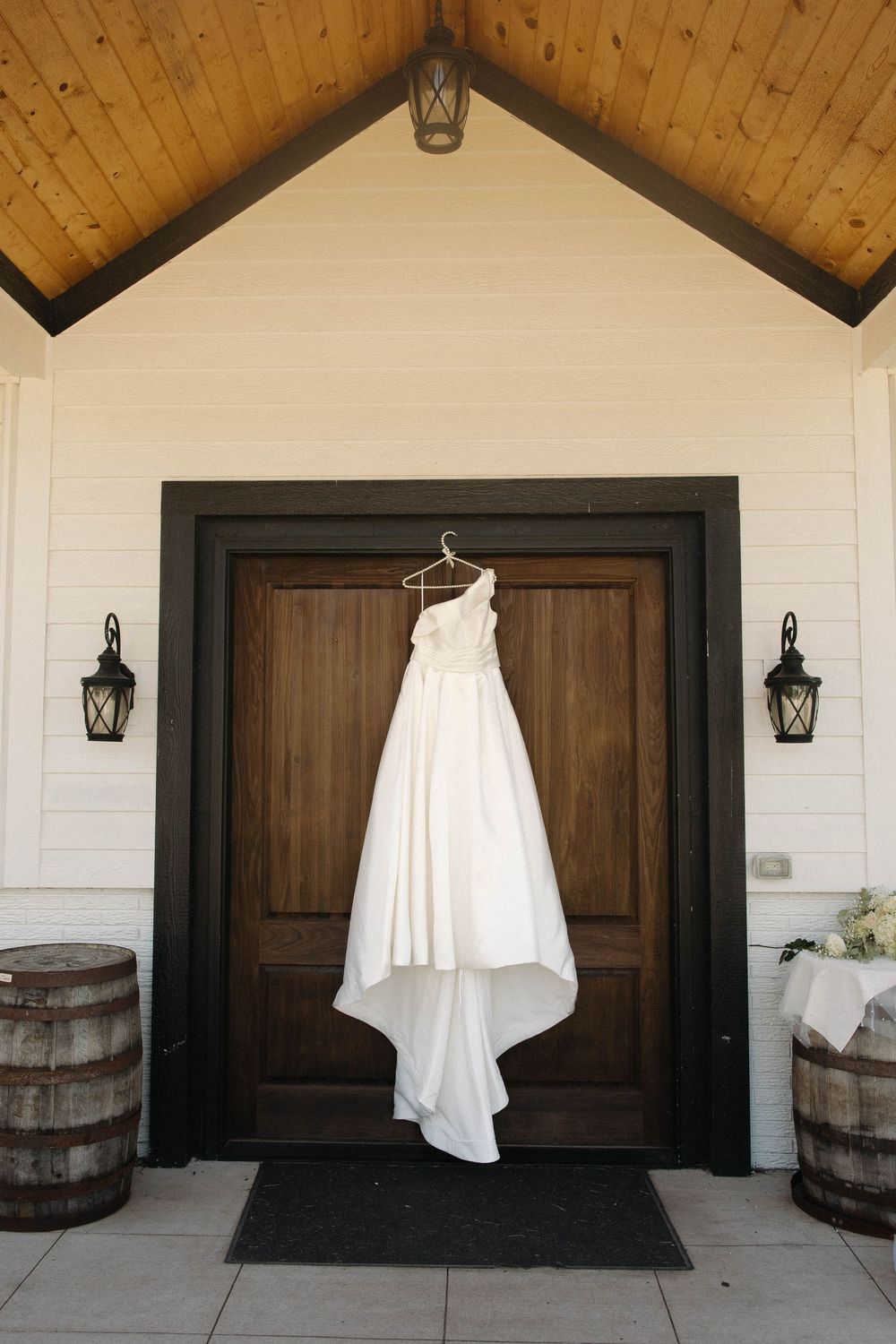 Wedding dress hanging outside white chapel doors with wooden barrels and rustic lanterns.