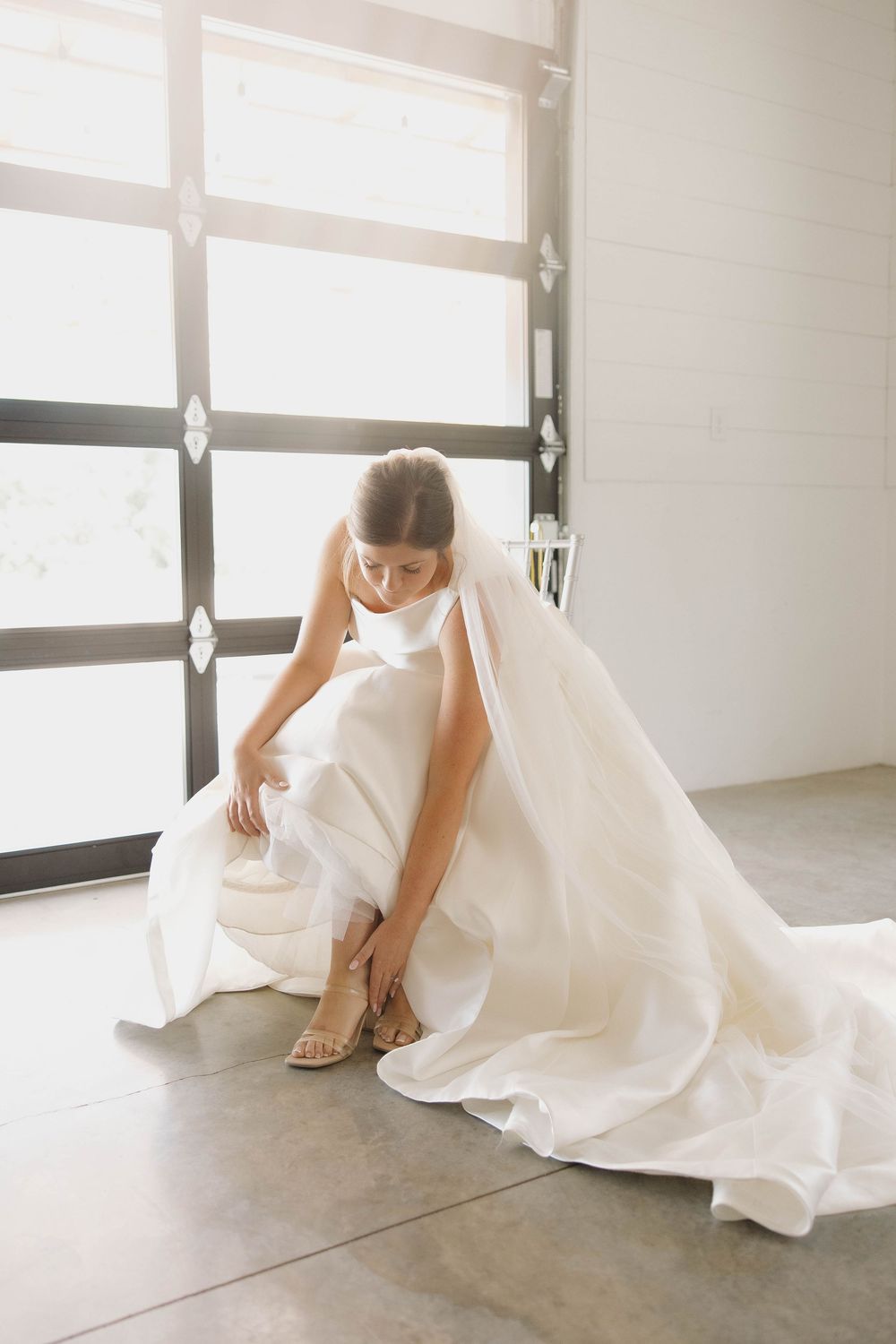 Bride in flowing white wedding dress adjusts her shoe in a minimalist indoor setting.