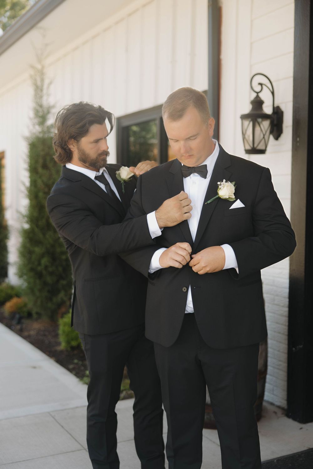 Groomsmen in black tuxedos share a lighthearted moment outside a white building during wedding day preparations.