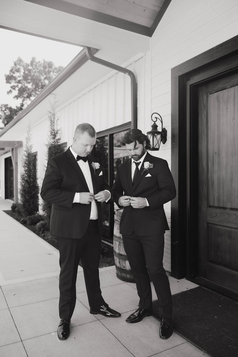 Groomsmen in black tuxedos share a lighthearted moment while reading outside.