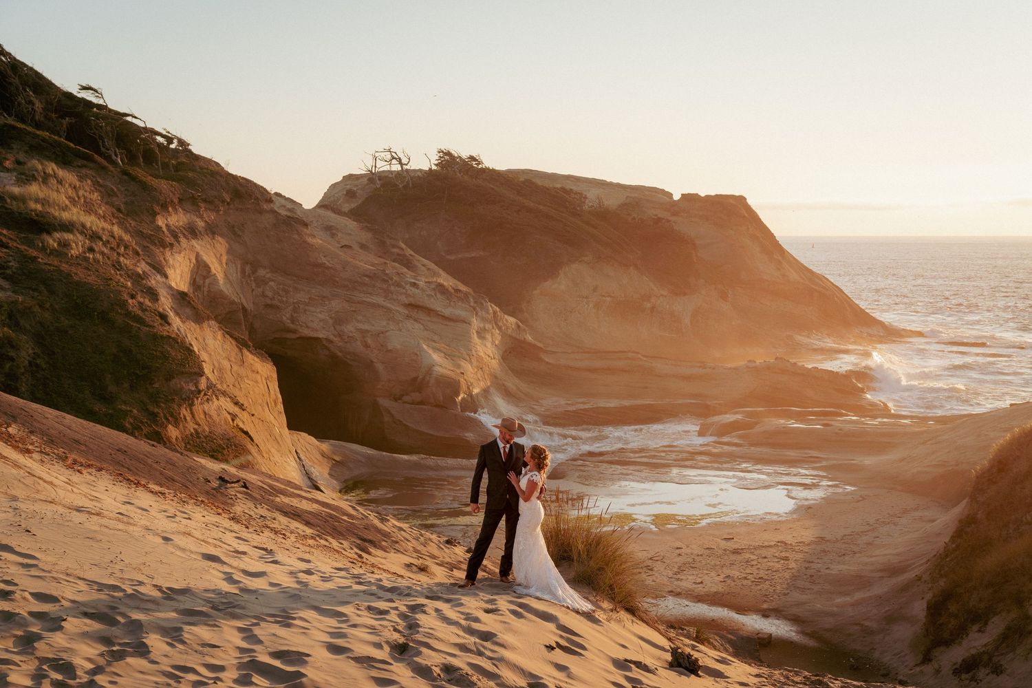 Bride and groom standing on a sandy cliff at sunset with ocean waves and rocky cliffs behind them at Cape Kiwanda, Oregon Coast.