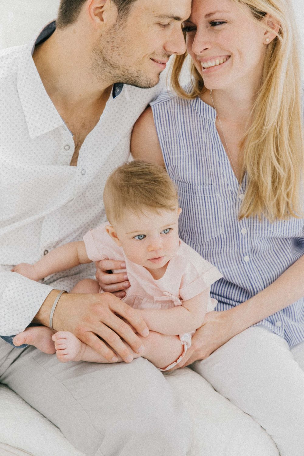 Tender family moment with parents and baby in casual attire on white bedding.Fotografia profissional de maternidade, recém-nascido e família