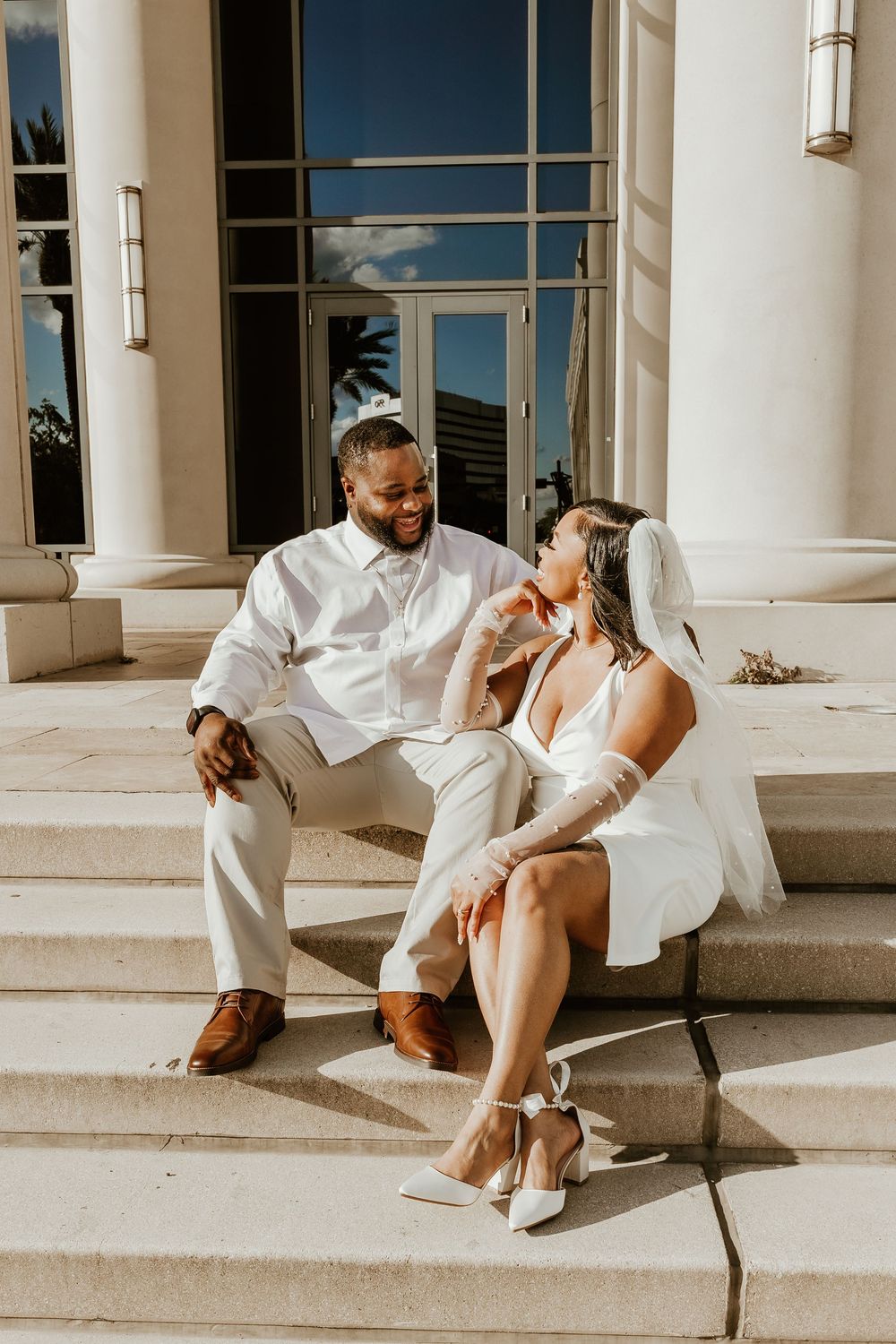 A couple in white wedding attire sits together on sunlit stone steps sharing a joyful moment.