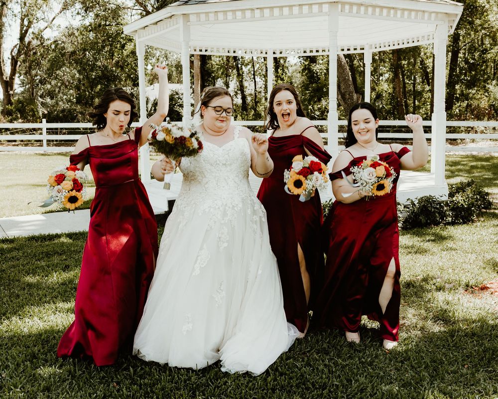 Wedding party in burgundy dresses and white gown pose outdoors on sunny day in garden setting.