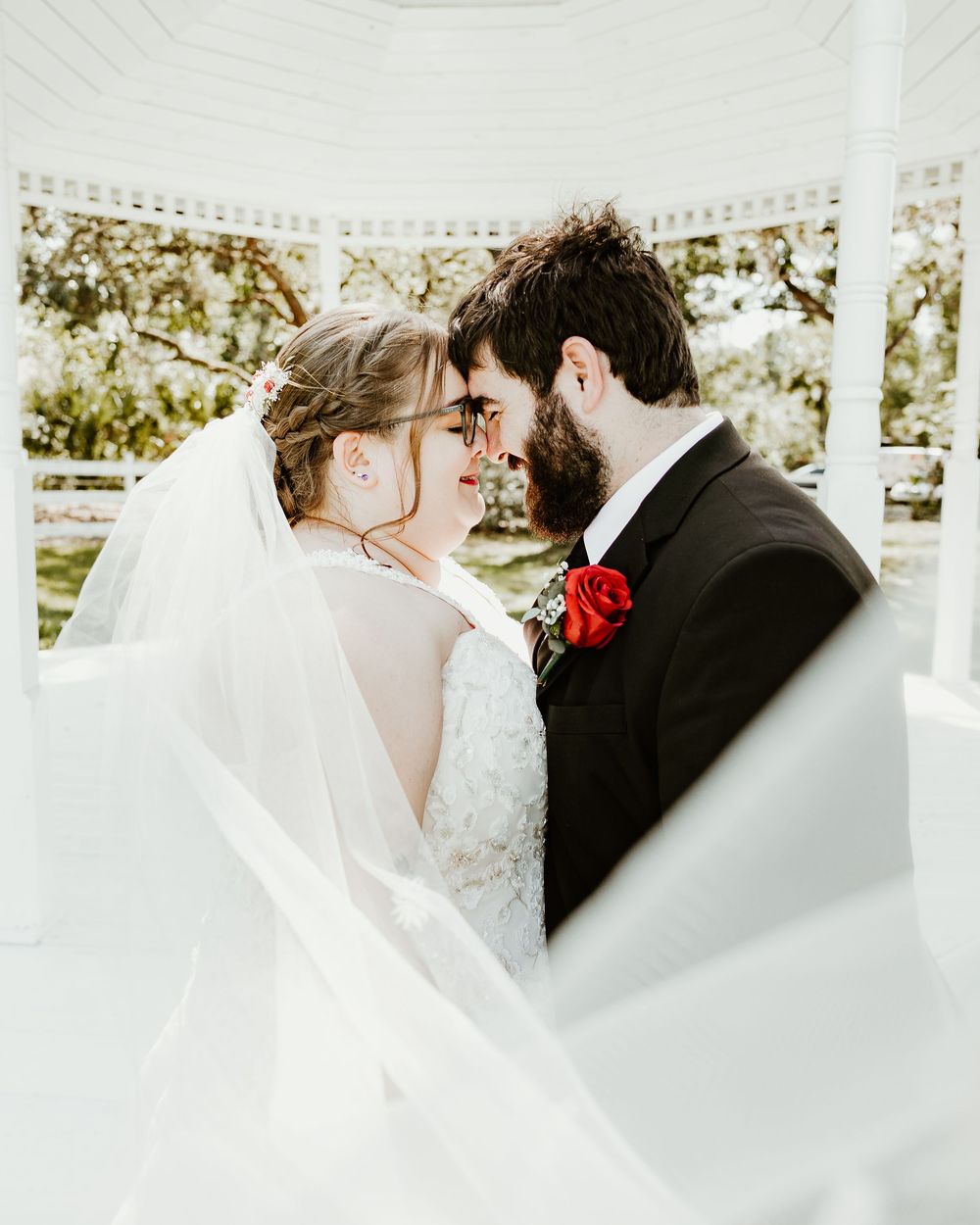 Intimate wedding portrait showing couple in formal attire with flowing white veil and red rose boutonniere under a gazebo.