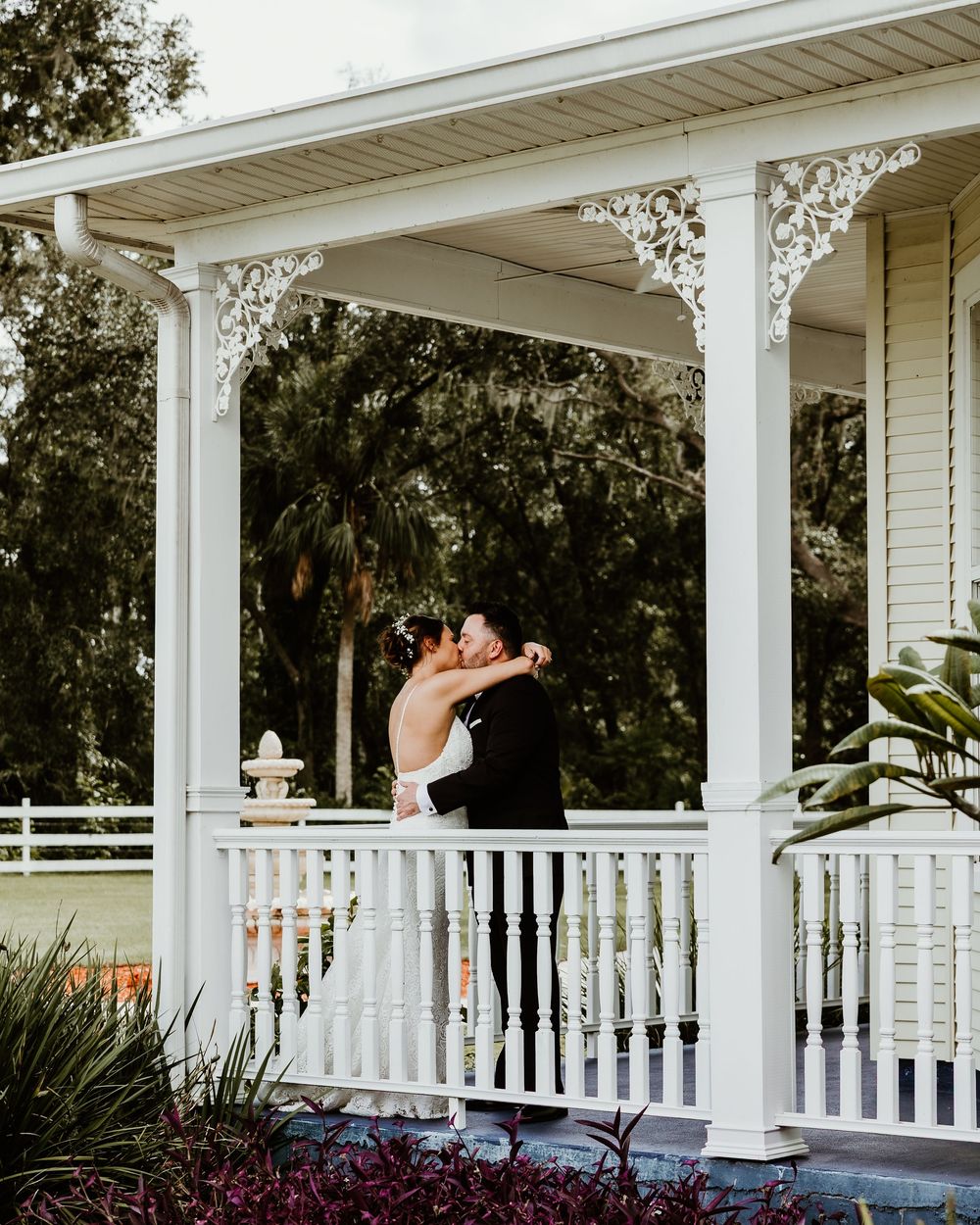 A couple embraces on a classic white wraparound porch with ornate columns and lush landscaping in the background.