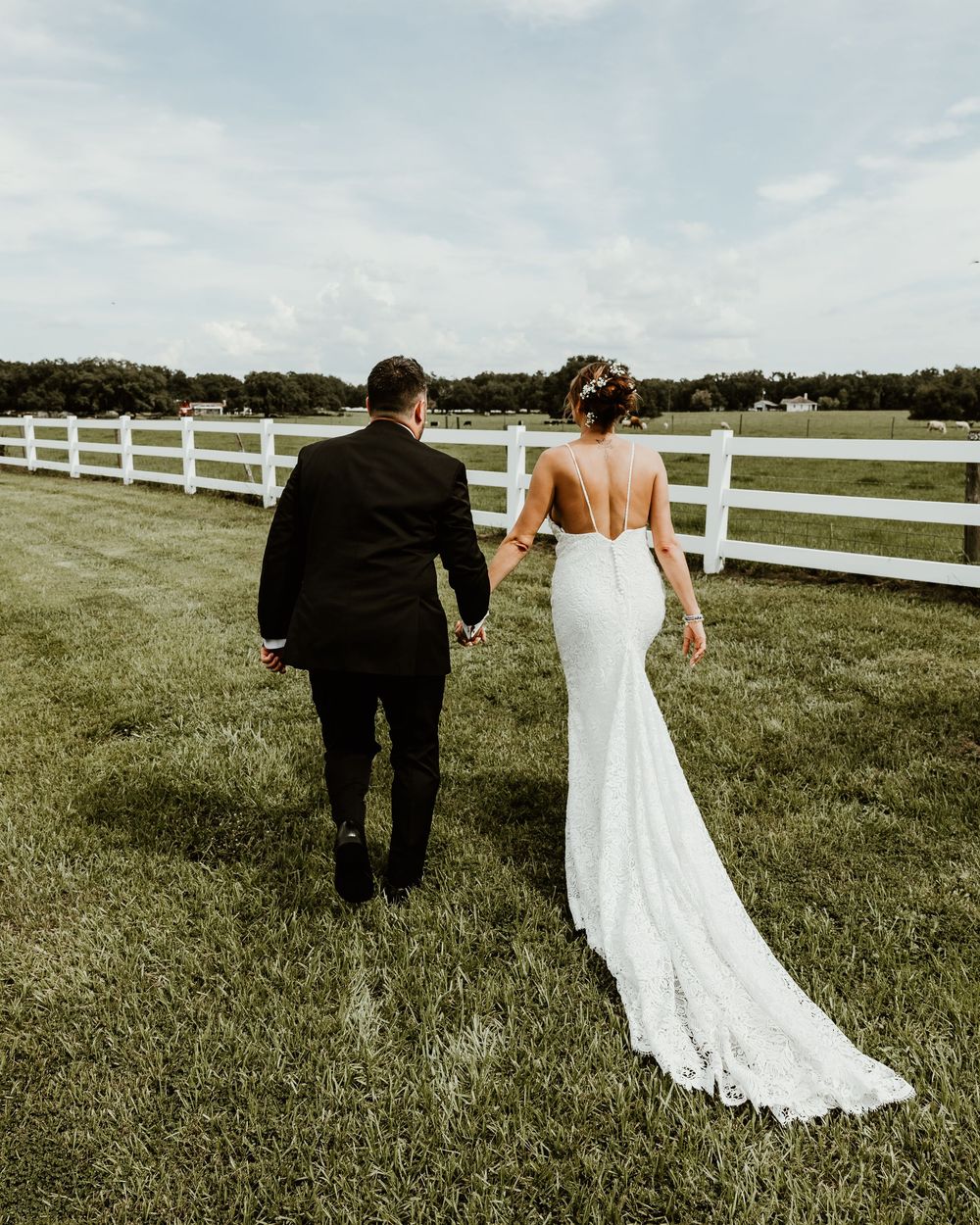 A couple in wedding attire walking across a scenic green field with white fences on a cloudy day.