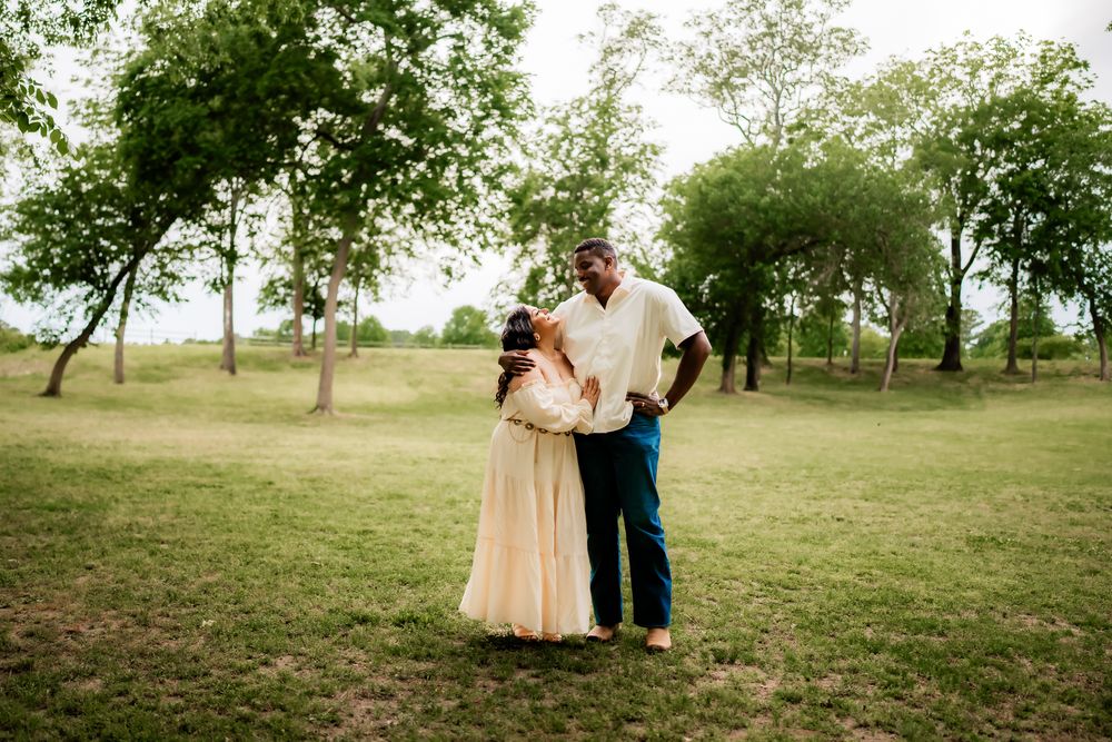 A romantic couple in a pastoral field setting wearing cream and blue outfits embrace during a photoshoot.