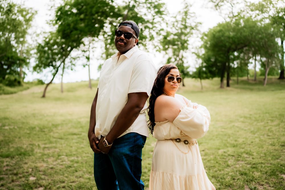 A stylish couple in sunglasses poses back-to-back in a grassy field wearing complementary outfits.