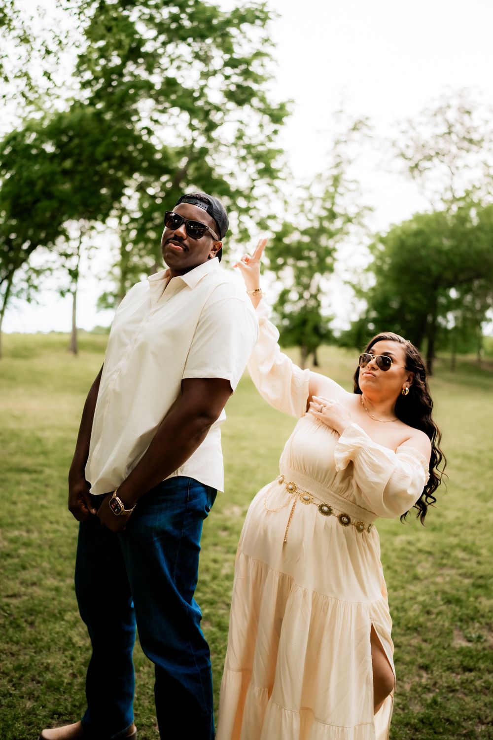 A couple dances together in a grassy field with trees on a sunny day, wearing coordinated cream and blue outfits.