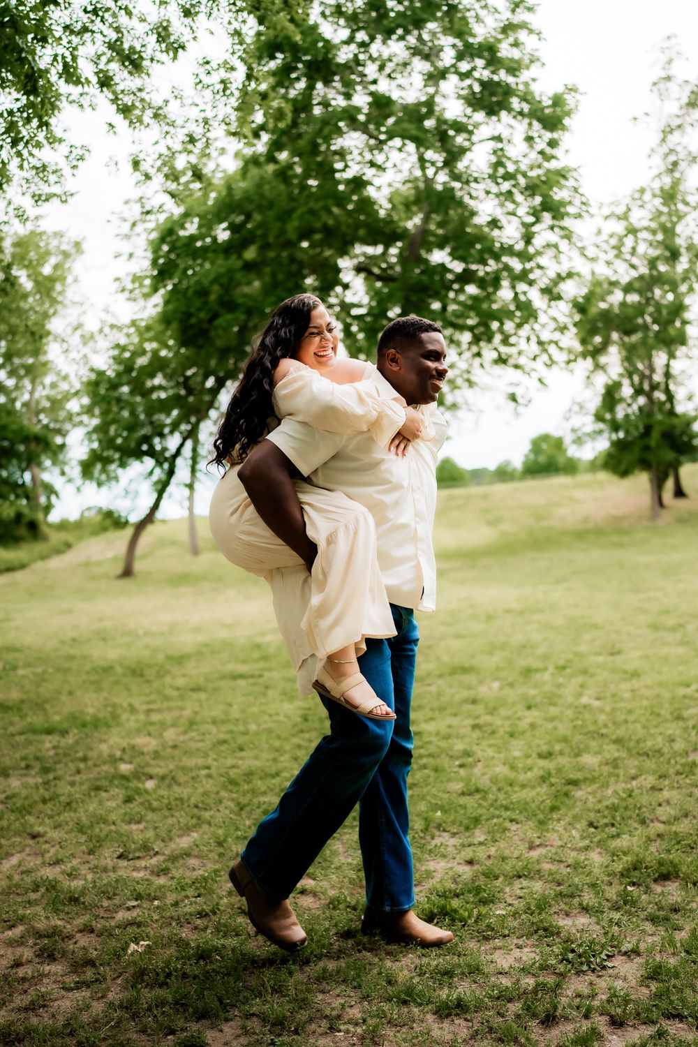 A playful couple shares a piggyback ride moment in a sunny field during their photoshoot.