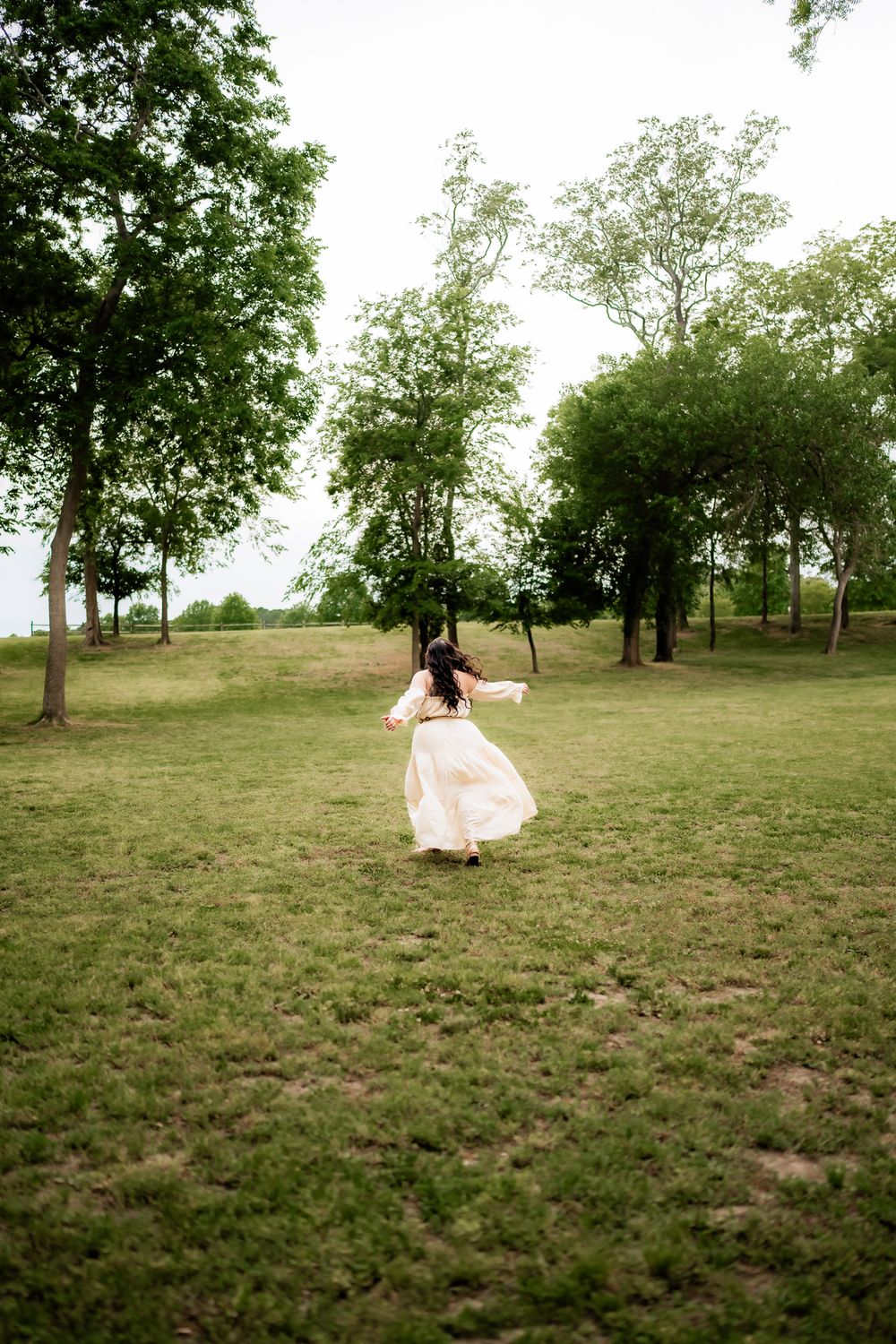 A person in a flowing cream dress dances and twirls freely in a pastoral field surrounded by trees.