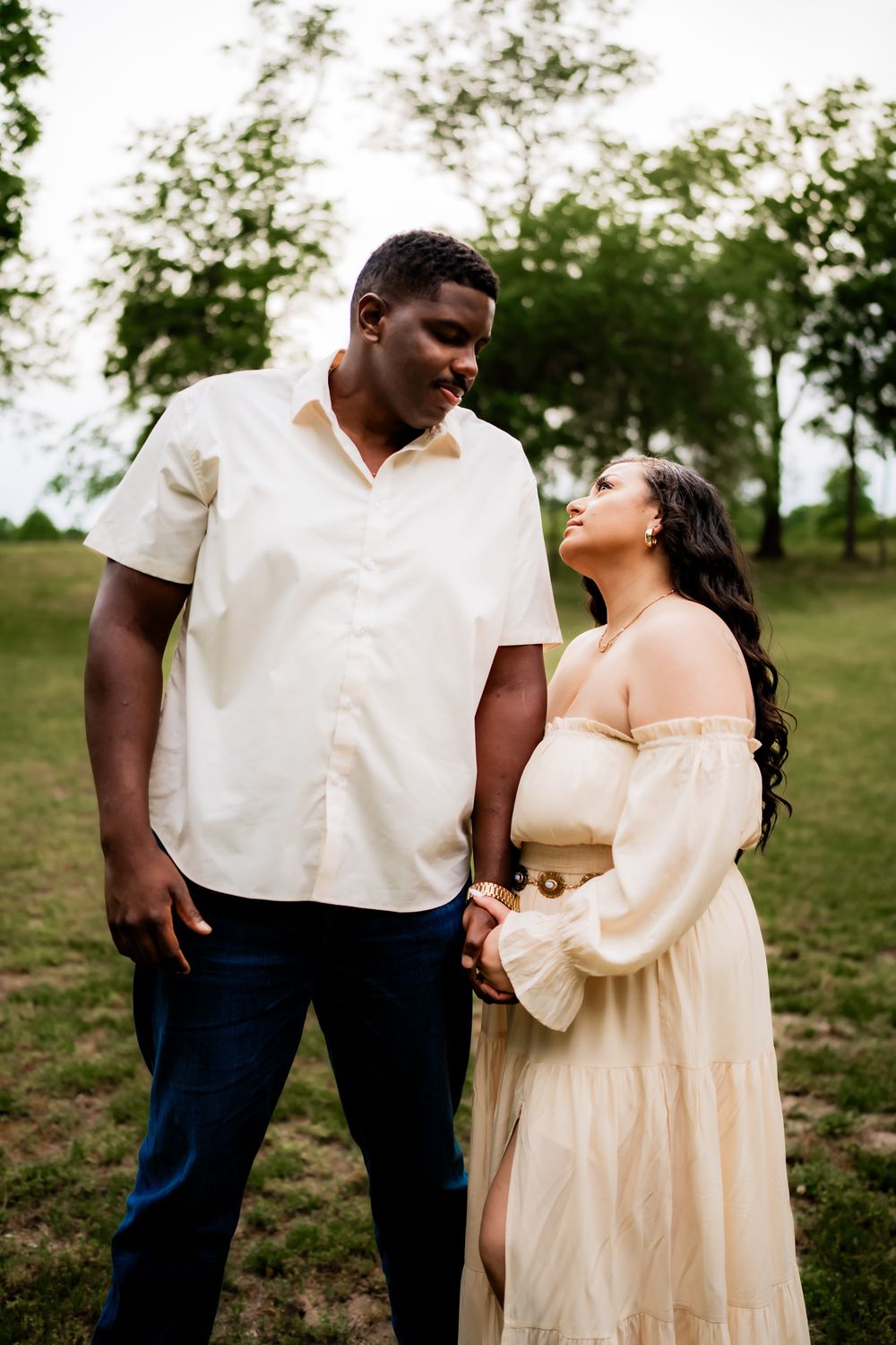 A couple stands lovingly together in a grassy outdoor setting, wearing white and cream colored outfits.