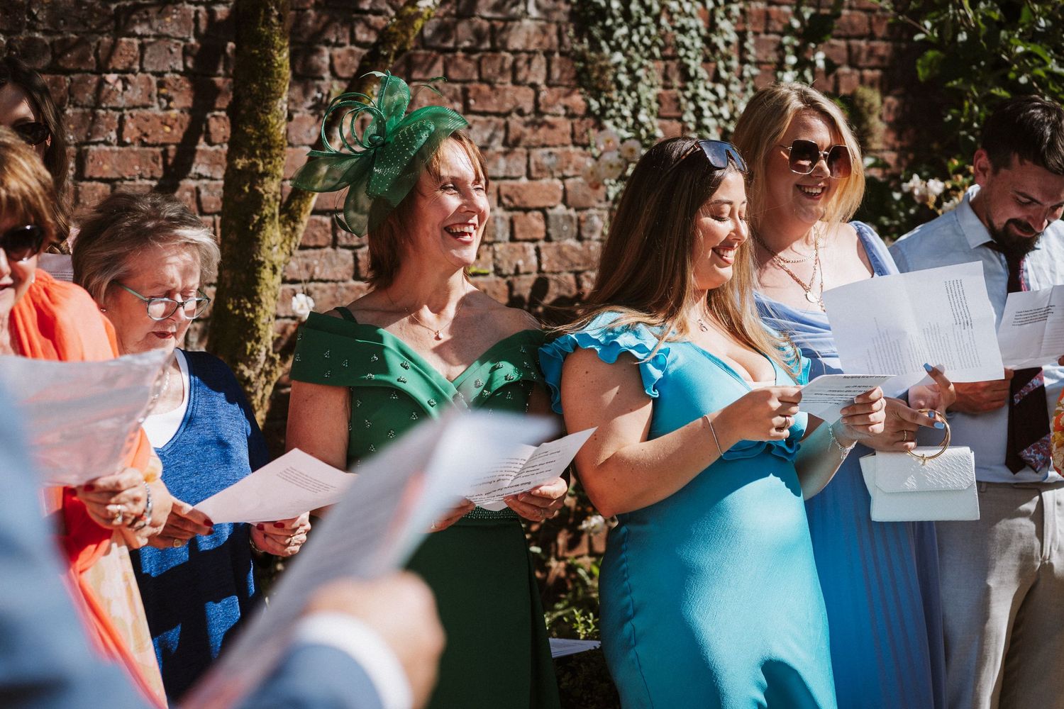 A group of wedding guests standing in a line against a brick wall hold up signs during an outdoor celebration.