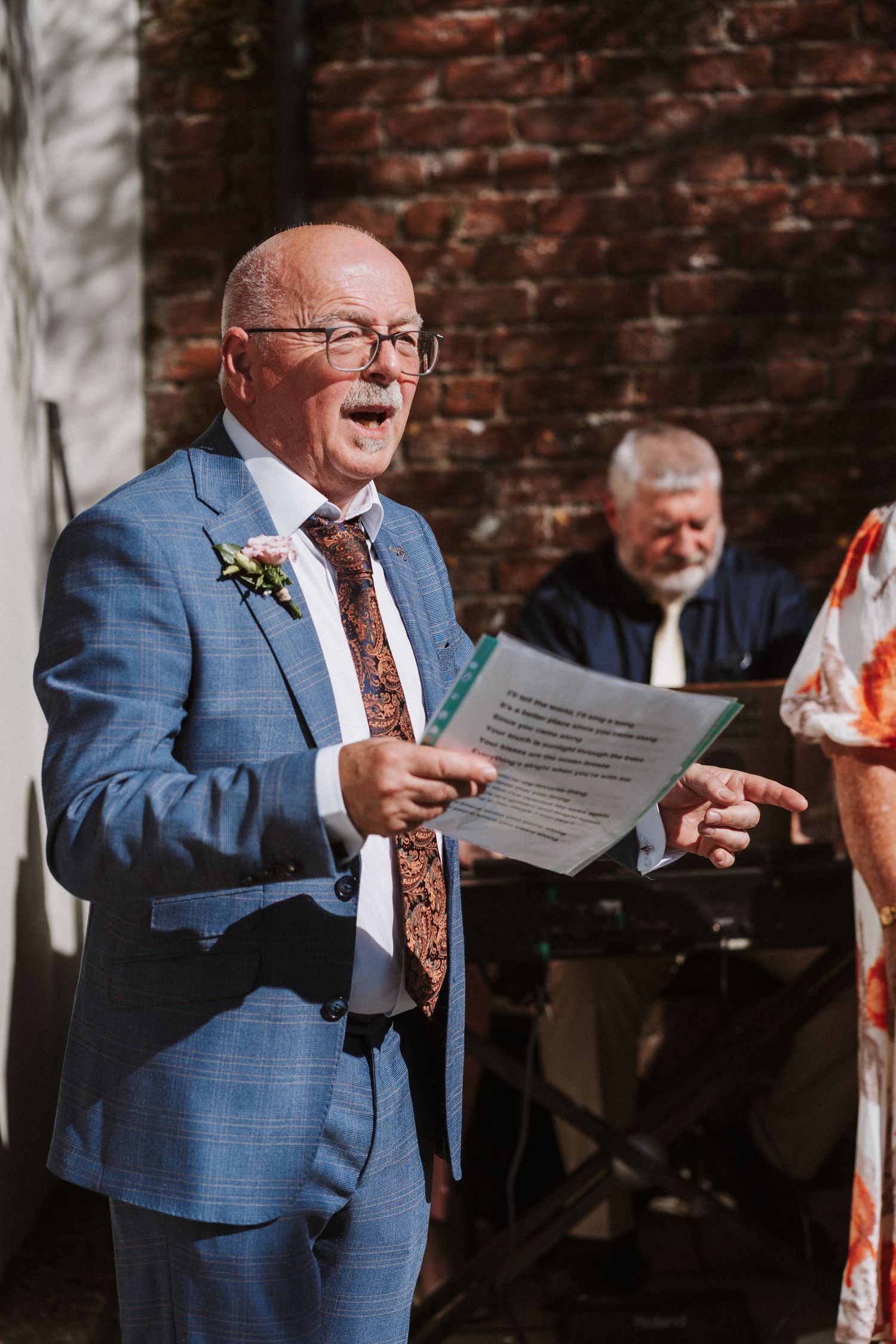 Man in blue suit and brown tie reads from paper document while standing by brick wall at outdoor event.