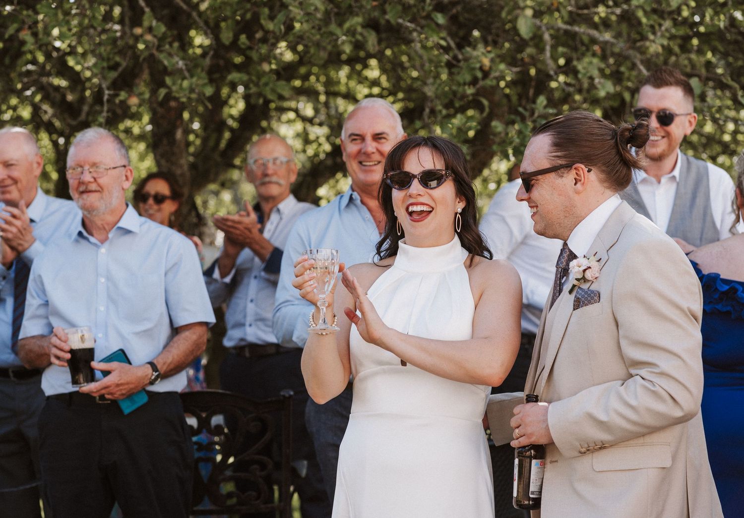 Wedding guests raise their glasses and smile during a toast at a sunny outdoor celebration.