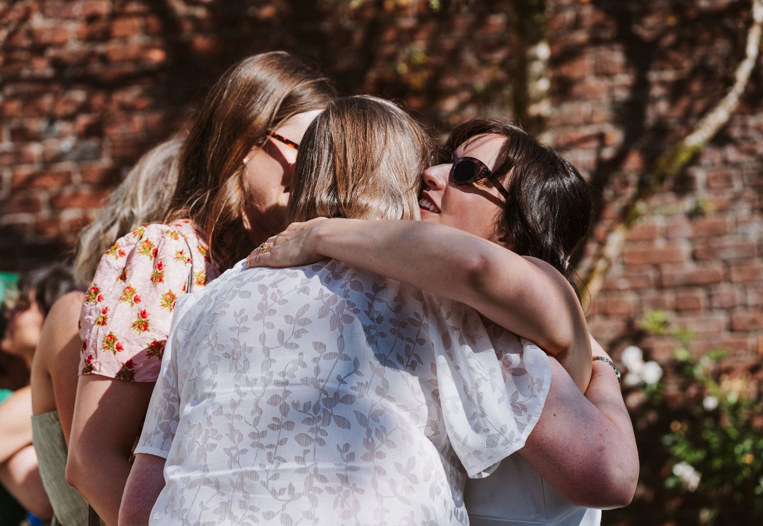 Wedding guests gather outdoors against a rustic brick wall backdrop during a sunny celebration.