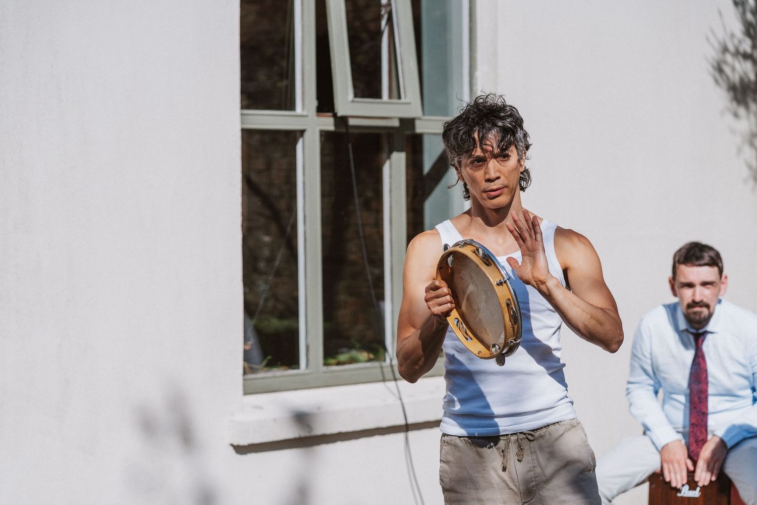 Two people stand outside a building, one wearing a tank top holding a boot while another wears a dress shirt and tie.