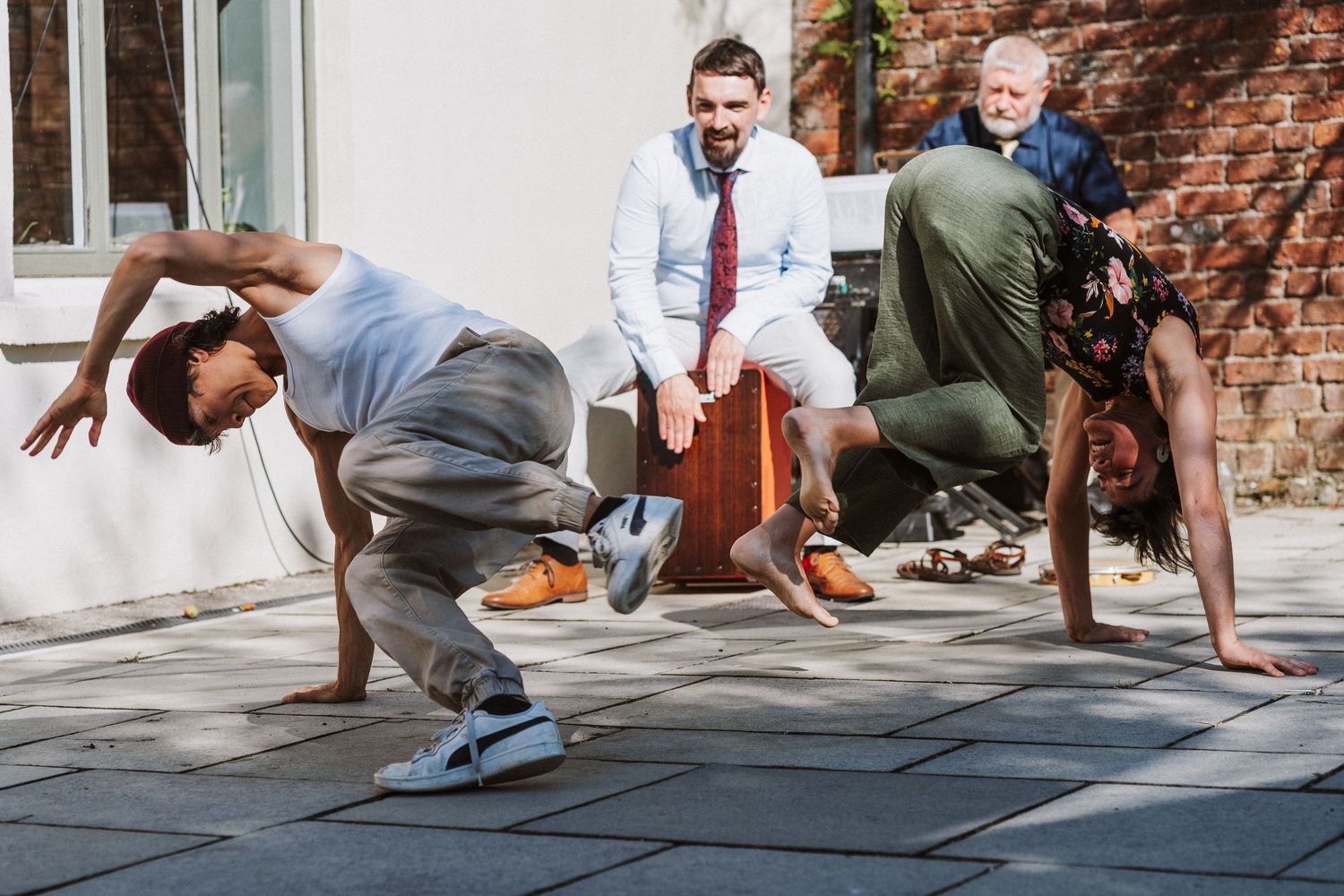 Three people practicing breakdancing moves on a stone paved walkway next to a brick wall.