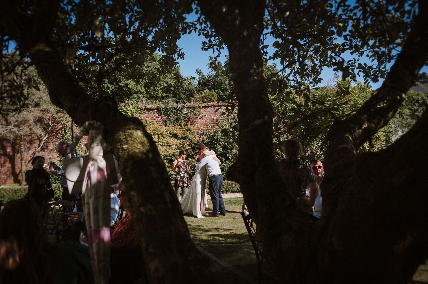 Guests gather outdoors under large shady trees during a summer celebration at a park.