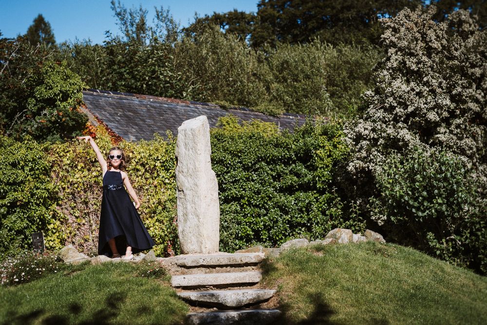 A scenic mountain trail leads through lush greenery to a moss-covered stone pillar in a sunlit garden setting.