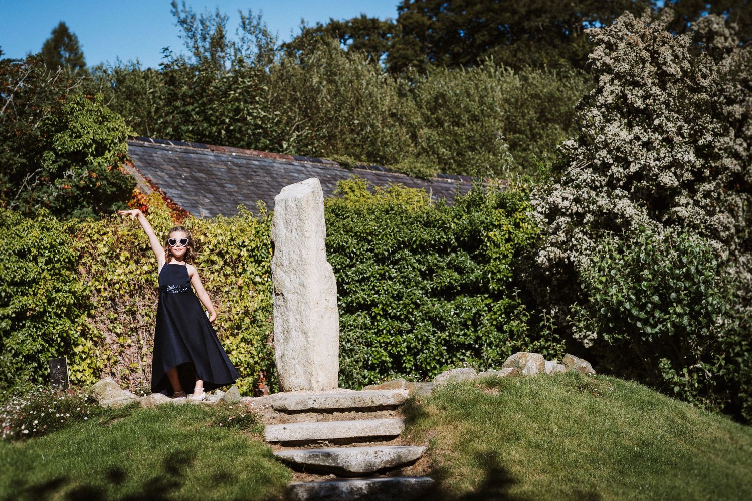 A scenic mountain trail leads through lush greenery to a moss-covered stone pillar in a sunlit garden setting.