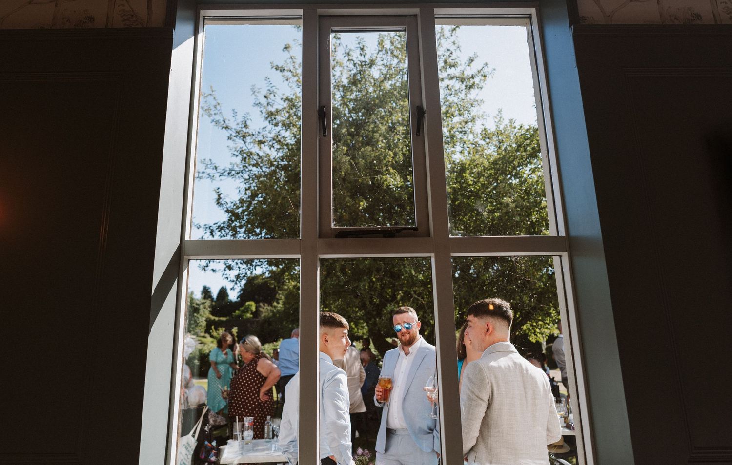A bride and groom stand together at their wedding ceremony while guests watch through large glass windows.