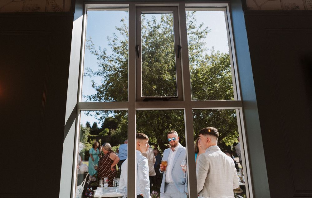 A bride and groom stand together at their wedding ceremony while guests watch through large glass windows.